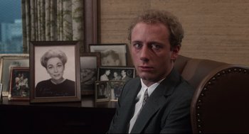 Movie still from “Mommie Dearest” (1981), directed by Frank Perry – A man in a suit sitting in front of a picture of a woman; Close Up shot, Over the shoulder angle