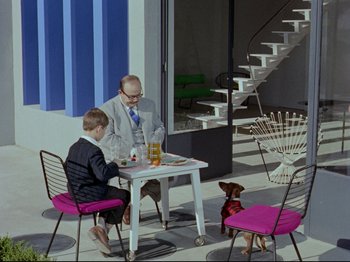 Movie still from “My Uncle” (1958), directed by Jacques Tati – A man sitting at a table with a dog; Wide shot, High angle
