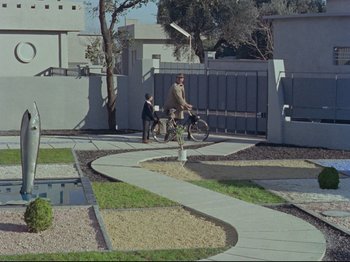 Movie still from “My Uncle” (1958), directed by Jacques Tati – A man riding a bike down a sidewalk next to trees; Extreme Wide shot, High angle