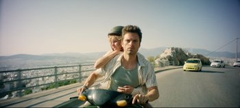 Movie still from “Monday” (2020), directed by Argyris Papadimitropoulos – A man and a woman riding a motorcycle down a street; Medium shot, Low angle