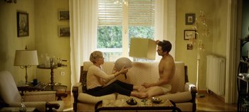 Movie still from “Monday” (2020), directed by Argyris Papadimitropoulos – A man and a woman sitting on a couch; Wide shot, Low angle