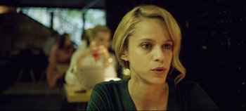 Movie still from “Monday” (2020), directed by Argyris Papadimitropoulos – A young woman sitting in front of a group of people; Close Up shot, Over the shoulder angle