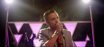 Movie still from “Monday” (2020), directed by Argyris Papadimitropoulos – A man in a suit is holding a microphone; Close Up shot, Low angle