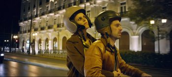 Movie still from “Monday” (2020), directed by Argyris Papadimitropoulos – A man and a woman riding a motorcycle down a street; Medium shot, Low angle