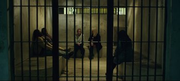 Movie still from “Monday” (2020), directed by Argyris Papadimitropoulos – Two people sitting in a jail cell with bars around them; Wide shot, High angle