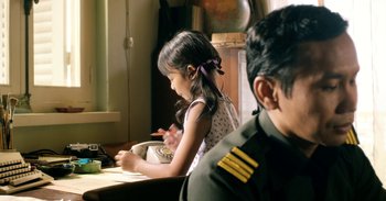 Movie still from “First They Killed My Father” (2017), directed by Angelina Jolie – A little girl sitting at a table in front of an officer; Medium shot, Over the shoulder angle