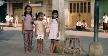 Movie still from “First They Killed My Father” (2017), directed by Angelina Jolie – Three young girls standing next to a wooden stool on the sidewalk; Wide shot, High angle