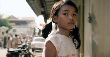 Movie still from “First They Killed My Father” (2017), directed by Angelina Jolie – A little girl with pigtails standing on the side of the street; Close Up shot, Over the shoulder angle
