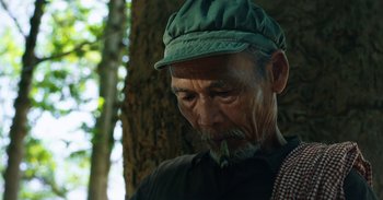 Movie still from “First They Killed My Father” (2017), directed by Angelina Jolie – An older man wearing a green hat smoking a cigarette; Close Up shot, Low angle