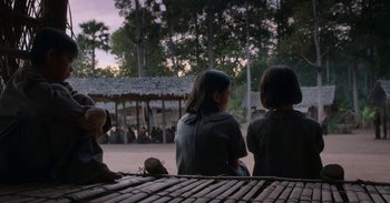 Movie still from “First They Killed My Father” (2017), directed by Angelina Jolie – Two women sitting on a bench in front of a thatched hut; Wide shot, Over the shoulder angle