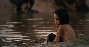 Movie still from “First They Killed My Father” (2017), directed by Angelina Jolie – A little girl sitting on the ground in a body of water; Medium shot, High angle