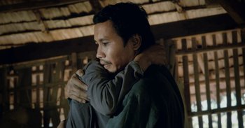 Movie still from “First They Killed My Father” (2017), directed by Angelina Jolie – A man is hugging another man in a hut; Medium shot, Over the shoulder angle