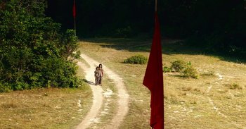 Movie still from “First They Killed My Father” (2017), directed by Angelina Jolie – Two people walking down a dirt road near trees and red flags; Extreme Wide shot, High angle