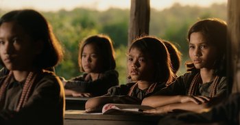 Movie still from “First They Killed My Father” (2017), directed by Angelina Jolie – A group of children sitting at a table with a book; Medium shot, Over the shoulder angle