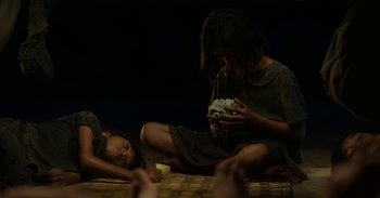 Movie still from “First They Killed My Father” (2017), directed by Angelina Jolie – A woman sitting on the ground with a child holding something; Medium shot, Over the shoulder angle