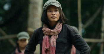 Movie still from “First They Killed My Father” (2017), directed by Angelina Jolie – A woman wearing a hat and a scarf is standing outside; Medium shot, Low angle
