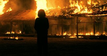 Movie still from “First They Killed My Father” (2017), directed by Angelina Jolie – A person standing in front of a burning building; Extreme Wide shot, Over the shoulder angle