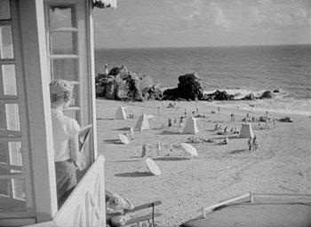 Movie still from “Monsieur Hulot's Holiday” (1953), directed by Jacques Tati – An old black and white photo of a woman looking out a window at the beach; Extreme Wide shot, High angle