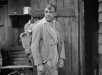 Movie still from “Monsieur Hulot's Holiday” (1953), directed by Jacques Tati – A man with a backpack standing in front of a wooden wall; Medium shot, Over the shoulder angle