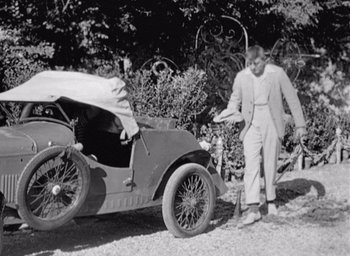 Movie still from “Monsieur Hulot's Holiday” (1953), directed by Jacques Tati – An old photo of a man standing next to an old car; Wide shot, High angle