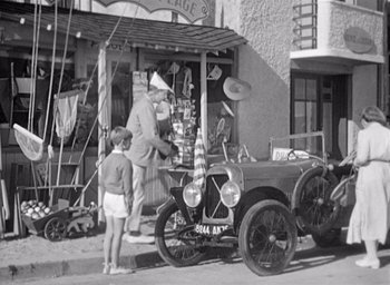 Movie still from “Monsieur Hulot's Holiday” (1953), directed by Jacques Tati – An old photo of a man standing next to an antique car; Wide shot, Over the shoulder angle