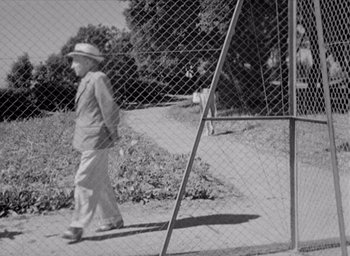 Movie still from “Monsieur Hulot's Holiday” (1953), directed by Jacques Tati – An older man walking down a sidewalk near a fence; Wide shot, Low angle