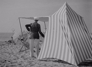 Movie still from “Monsieur Hulot's Holiday” (1953), directed by Jacques Tati – An old photo of a man standing on the beach with an awning; Wide shot, High angle