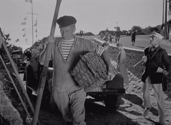 Movie still from “Monsieur Hulot's Holiday” (1953), directed by Jacques Tati – An old black and white photo of a man carrying a basket; Wide shot, Low angle