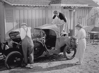 Movie still from “Monsieur Hulot's Holiday” (1953), directed by Jacques Tati – An old photo of a man and two women standing next to an old car; Wide shot, Low angle