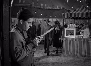 Movie still from “Moonrise” (1948), directed by Frank Borzage – A man holding a baseball bat while standing next to a tent; Medium shot, Over the shoulder angle