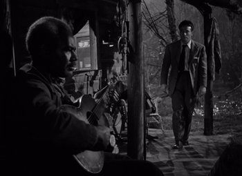 Movie still from “Moonrise” (1948), directed by Frank Borzage – A man playing a violin while another man walks by; Wide shot, Over the shoulder angle