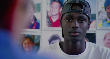 Movie still from “Morning Light” (2008), directed by Mark Monroe – A young man in a cap is in front of pictures; Close Up shot, Over the shoulder angle