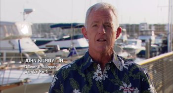 Movie still from “Morning Light” (2008), directed by Mark Monroe – An older man wearing a hawaiian shirt standing in front of boats; Medium shot, Low angle