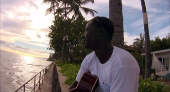 Movie still from “Morning Light” (2008), directed by Mark Monroe – A man is playing a guitar on the side of the beach; Medium shot, Low angle