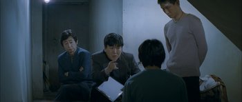 Movie still from “Mother” (2009), directed by Bong Joon Ho – A group of people sitting around a room; Medium shot, Over the shoulder angle