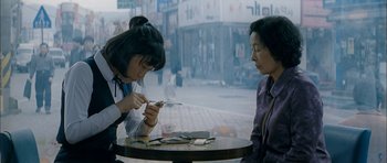 Movie still from “Mother” (2009), directed by Bong Joon Ho – Two women sitting at a table looking at their cell phones; Medium shot, High angle