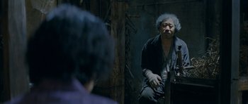 Movie still from “Mother” (2009), directed by Bong Joon Ho – An older man sitting on the ground in front of a wall; Medium shot, Over the shoulder angle