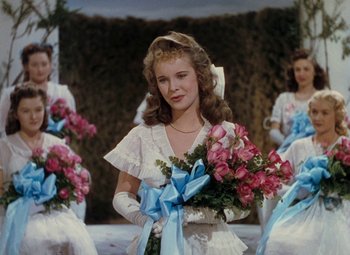 Movie still from “Mother Wore Tights” (1947), directed by Walter Lang – A woman in a white dress holding a bunch of pink flowers; Medium shot, Low angle