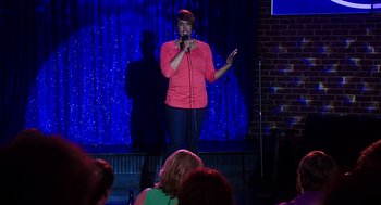 Movie still from “Mother's Day” (2016), directed by Garry Marshall – A woman standing on a stage holding a microphone; Wide shot, Over the shoulder angle