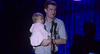 Movie still from “Mother's Day” (2016), directed by Garry Marshall – A man holding a baby while standing at a microphone; Medium shot, Over the shoulder angle