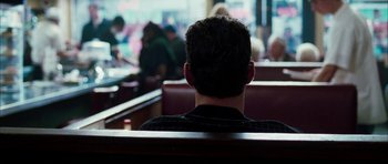 Movie still from “Mr. & Mrs. Smith” (2005), directed by Doug Liman – A man sitting on a bench in front of a group of people; Medium shot, Over the shoulder angle