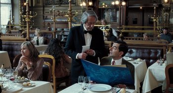 Movie still from “Mr. Bean's Holiday” (2007), directed by Steve Bendelack – Two men in suits are standing at a table; Medium shot, Over the shoulder angle