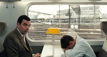 Movie still from “Mr. Bean's Holiday” (2007), directed by Steve Bendelack – A man reading a book on a train while another man sleeps on the train; Medium shot, High angle