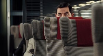Movie still from “Mr. Bean's Holiday” (2007), directed by Steve Bendelack – A man sitting on a train looking over the back of a seat; Medium shot, Over the shoulder angle