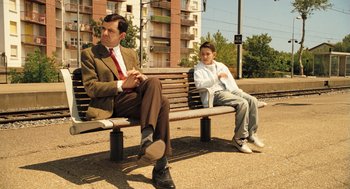 Movie still from “Mr. Bean's Holiday” (2007), directed by Steve Bendelack – A man sitting on a bench next to another man; Medium shot, Over the shoulder angle