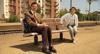 Movie still from “Mr. Bean's Holiday” (2007), directed by Steve Bendelack – A man and a woman sitting on a wooden bench; Medium shot, Low angle