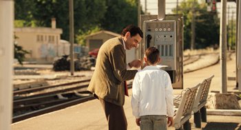 Movie still from “Mr. Bean's Holiday” (2007), directed by Steve Bendelack – A man and a boy standing next to each other on a train platform; Medium shot, Over the shoulder angle