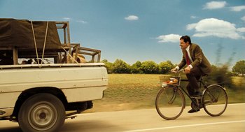 Movie still from “Mr. Bean's Holiday” (2007), directed by Steve Bendelack – A man riding a bike next to a white truck; Wide shot, Low angle