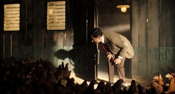 Movie still from “Mr. Bean's Holiday” (2007), directed by Steve Bendelack – A man in a suit and tie standing in front of a bunch of chickens; Medium shot, Low angle