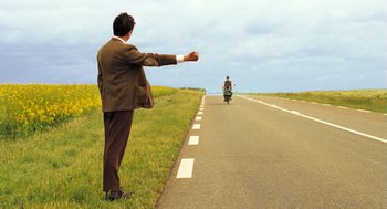 Movie still from “Mr. Bean's Holiday” (2007), directed by Steve Bendelack – A man standing on the side of a road directing a motorcycle; Wide shot, High angle